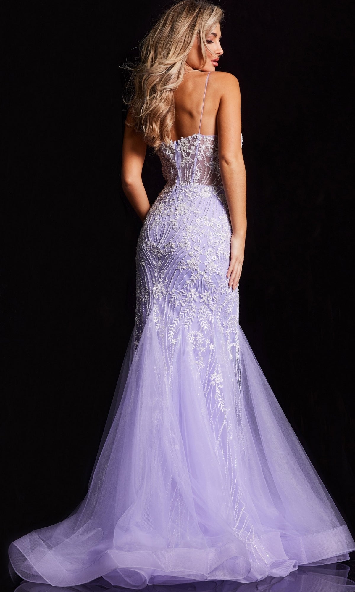 A woman with long blonde hair models the Sheer-Corset Long Mermaid Prom Dress (Jovani 37414) in lavender, featuring embroidery and a tulle train, shown from the back against a black background.